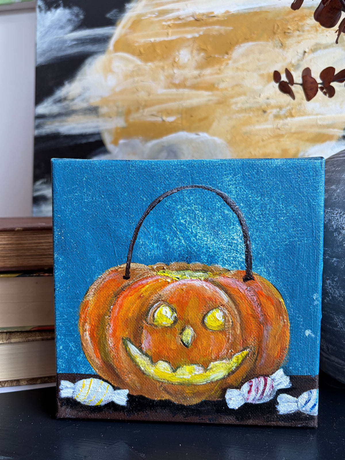 A small square canvas painting of a smiling jack-o'-lantern pumpkin with a handle, set against a blue background with candy and a dark base, displayed on a tabletop in front of a larger painting.