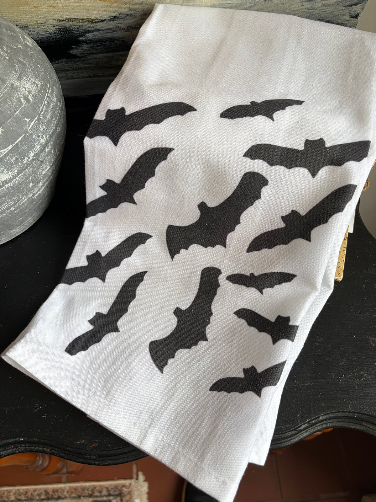 A white fabric napkin or tea towel printed with multiple black bat silhouettes, laid on a dark wooden surface next to a textured gray decorative object.