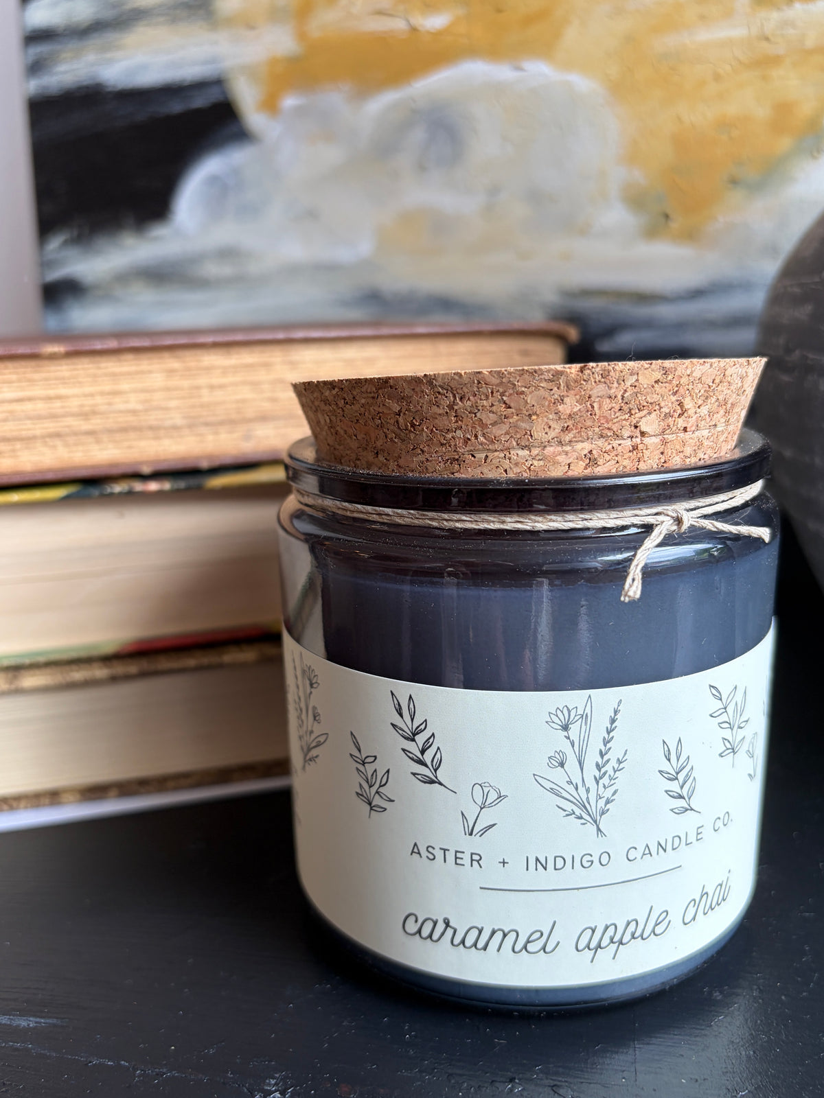 A glass jar candle with a dark charcoal wax body, a cork lid, and a cream label reading 'ASTER + INDIGO CANDLE CO' and 'caramel apple chai'. It sits on a dark tabletop with stacked books and a blurred painting in the background.