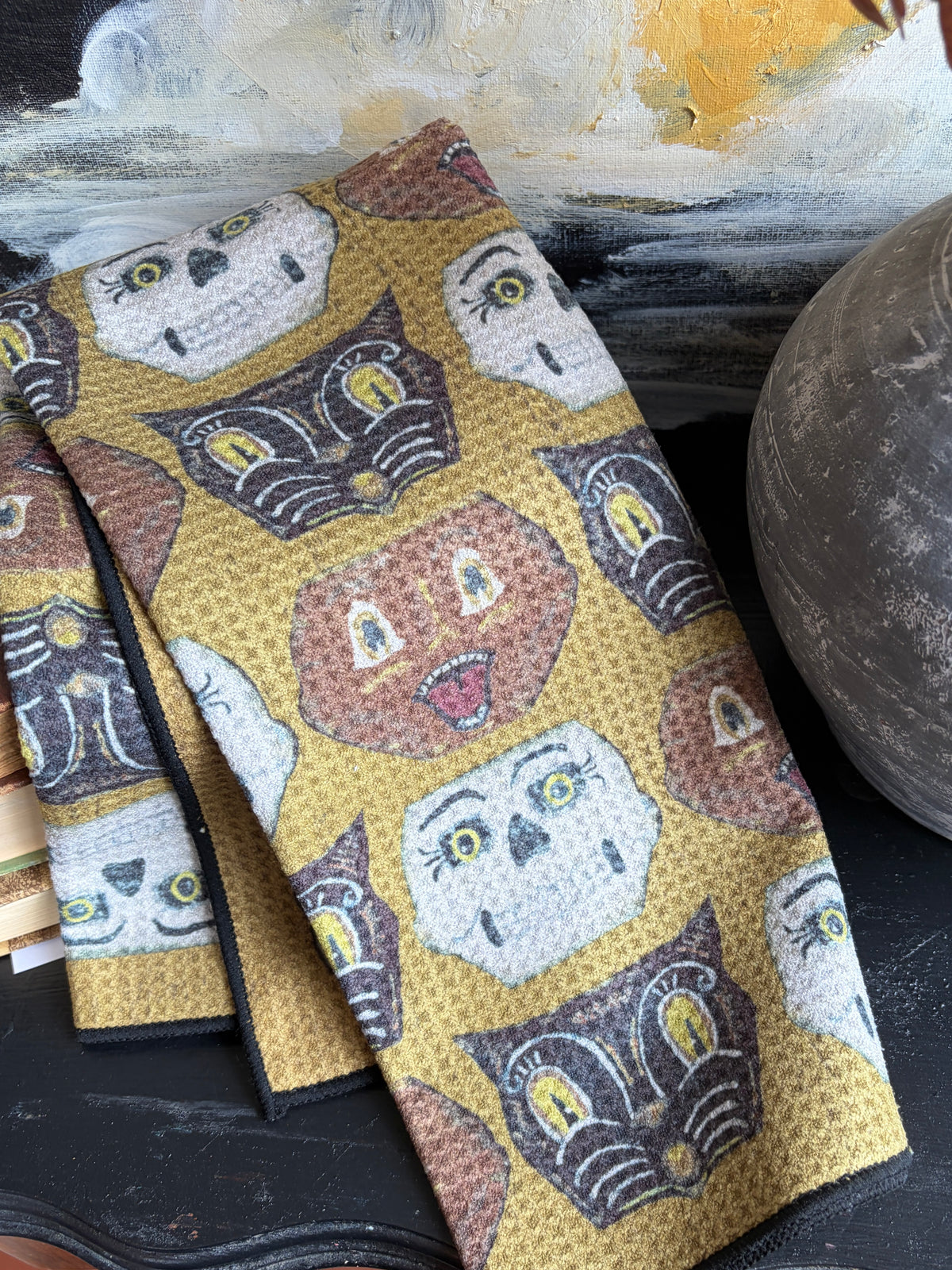 A folded fabric item, likely a napkin or small towel, featuring a repeating pattern of stylized owl faces in cream, brown, and dark gray on a mustard yellow background. The fabric is styled on a dark tabletop next to a textured ceramic vase and a painted canvas.