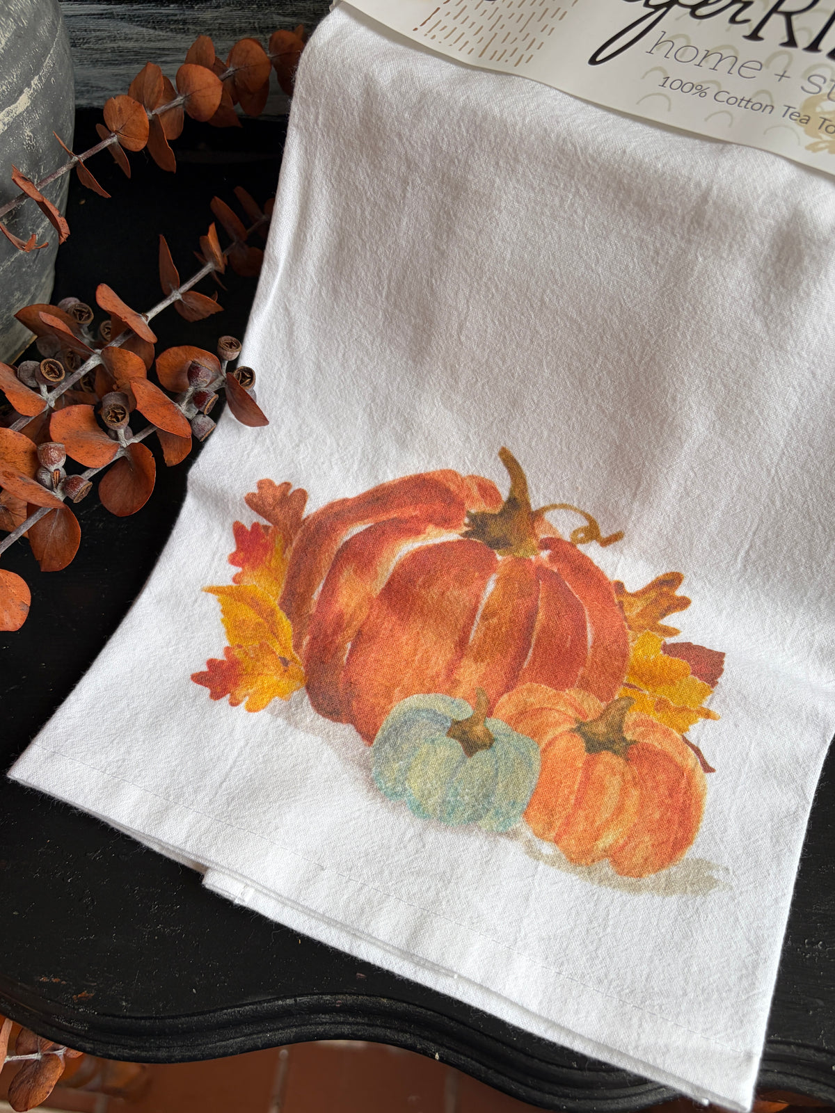 A white cotton tea towel with a watercolor-style autumn pumpkin print, laid flat on a dark wooden surface with decorative dried leaves and a paper header card partially visible at the top.