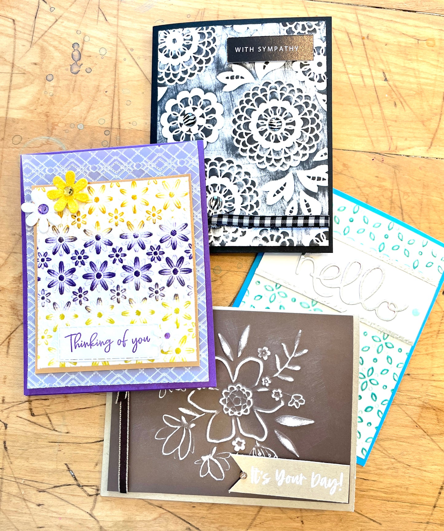Four greeting cards with floral designs on a wooden surface. It shows a set of embossed greeting cards. One says hello, others say with sympathy, it's your day and thinking of you with a yellow flower.