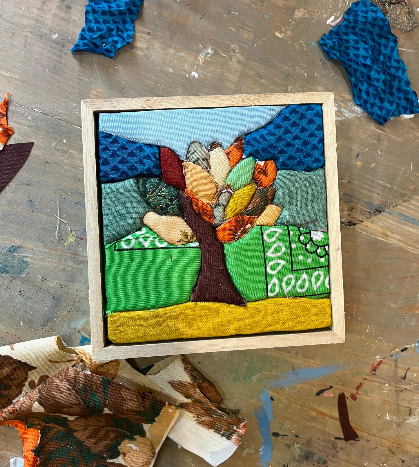 Textile art piece with a tree design on a wooden frame, surrounded by fabric scraps.