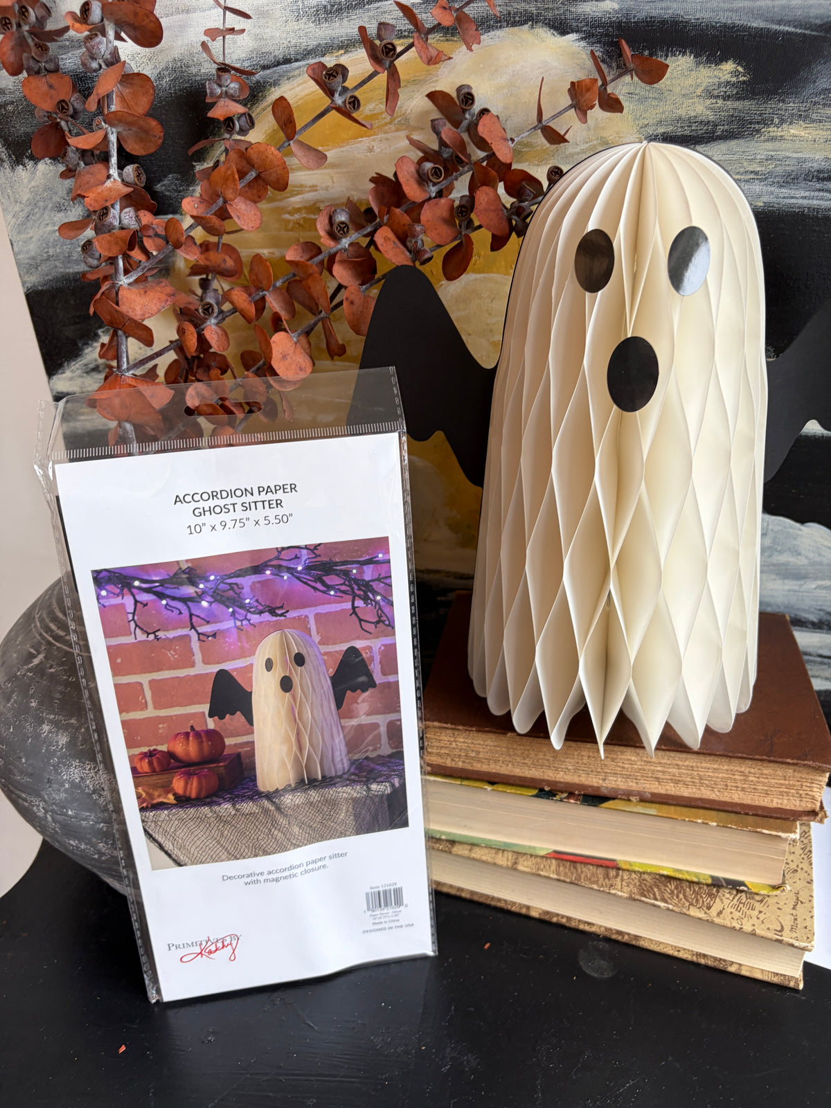 A packaged ghost-shaped paper ornament kit labeled 'Accordion Paper Ghost Set' stands beside a finished cream-colored accordion paper ghost decoration with black eyes and mouth, displayed on stacked books against a decorative autumn background.
