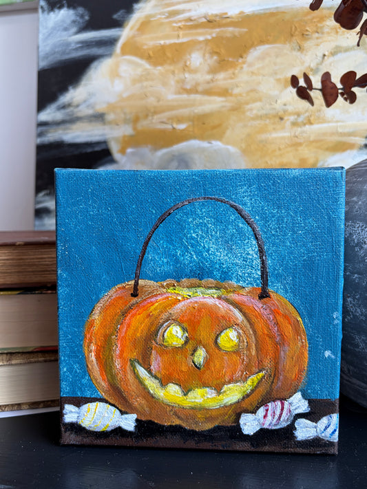 A small square canvas painting of a smiling jack-o'-lantern pumpkin with a handle, set against a blue background with candy and a dark base, displayed on a tabletop in front of a larger painting.
