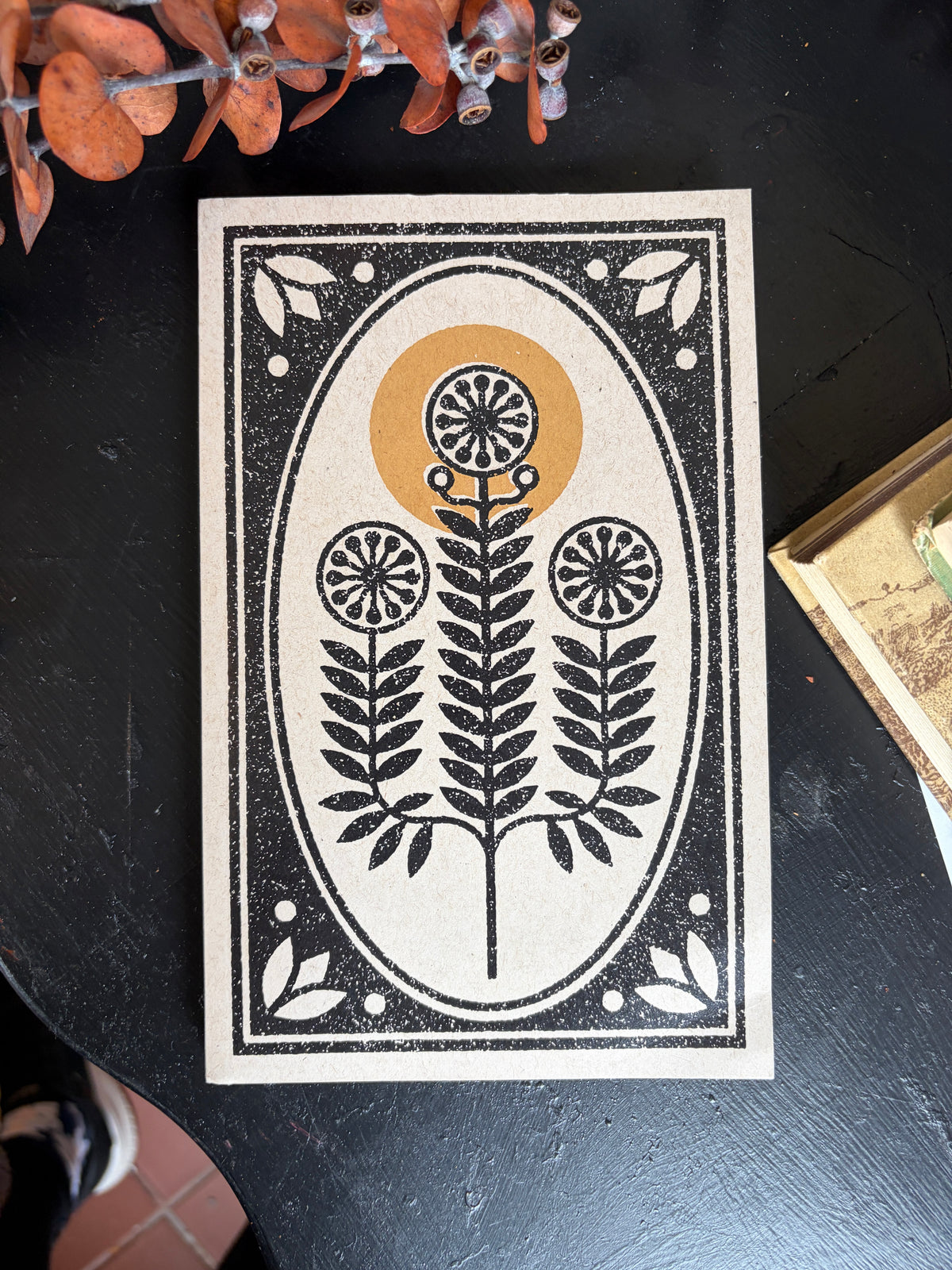 A rectangular decorative tile or print featuring a black botanical oval design on a cream background, placed on a dark tabletop with dried orange leaves and a small tray partially visible around it.