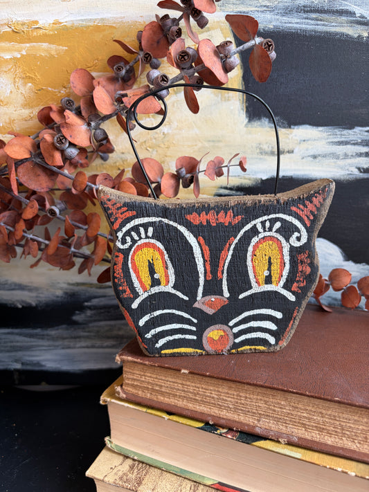 A hand-painted wooden cat-shaped basket with a black handle, resting on a stack of vintage books. The basket features a black base with white, orange, and yellow painted details.