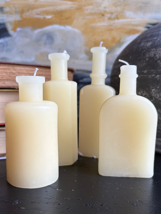Four handmade wax candles in bottle-shaped glass vessels with white wicks, arranged on a dark tabletop with a blurred rustic background.