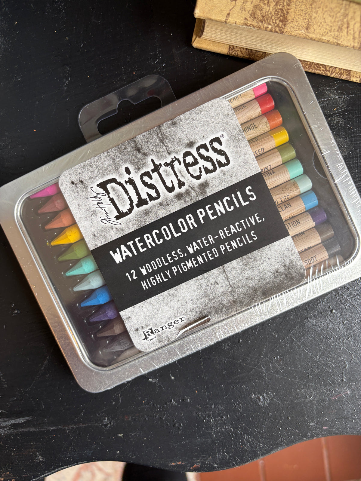 A 12-piece set of Ranger Distress Watercolor Pencils in a silver metal tin, displayed on a dark textured surface with a book in the background.