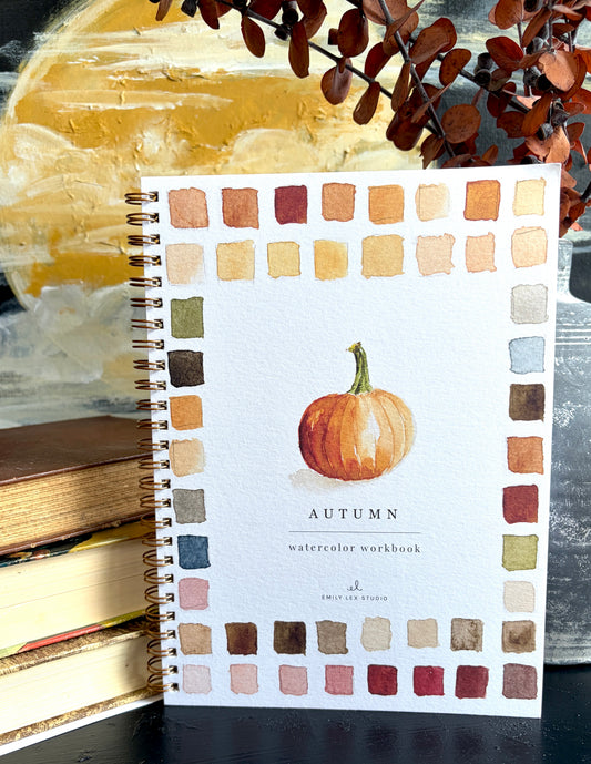 A spiral-bound watercolor workbook titled 'AUTUMN' with a watercolor pumpkin illustration and a border of autumn color swatches, styled on a dark surface with stacked books and dried leaves in the background.