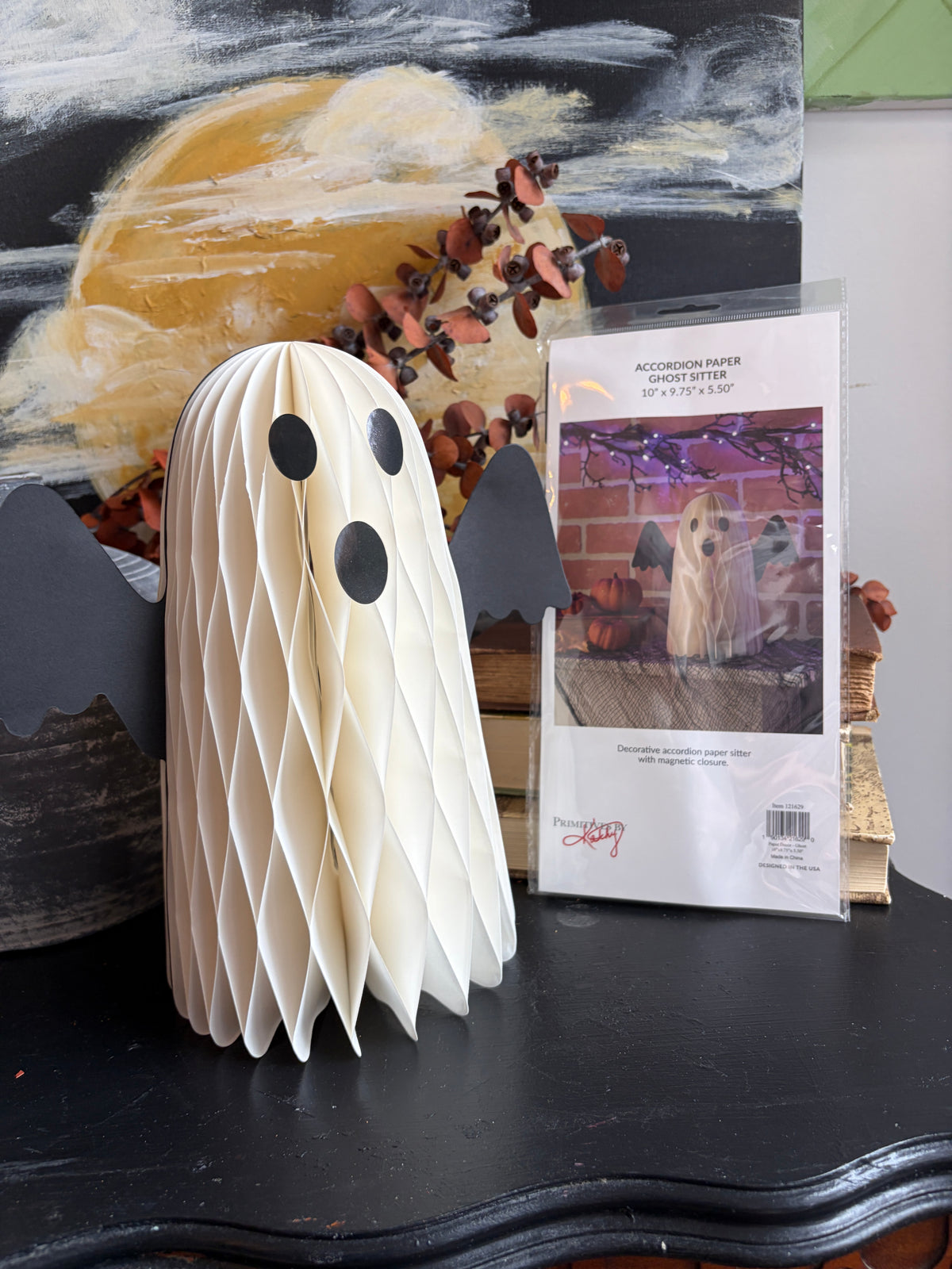 A cream-colored honeycomb paper ghost decoration with black eyes and black bat wings stands on a black table next to its retail packaging. The background features a colorful abstract painting and rustic autumn foliage.