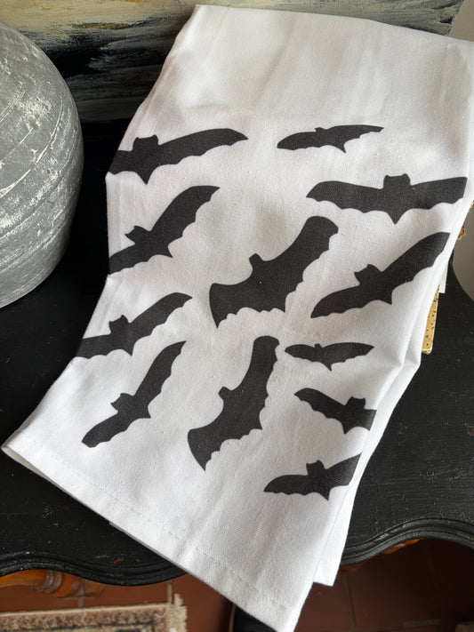 A white fabric napkin or tea towel printed with multiple black bat silhouettes, laid on a dark wooden surface next to a textured gray decorative object.
