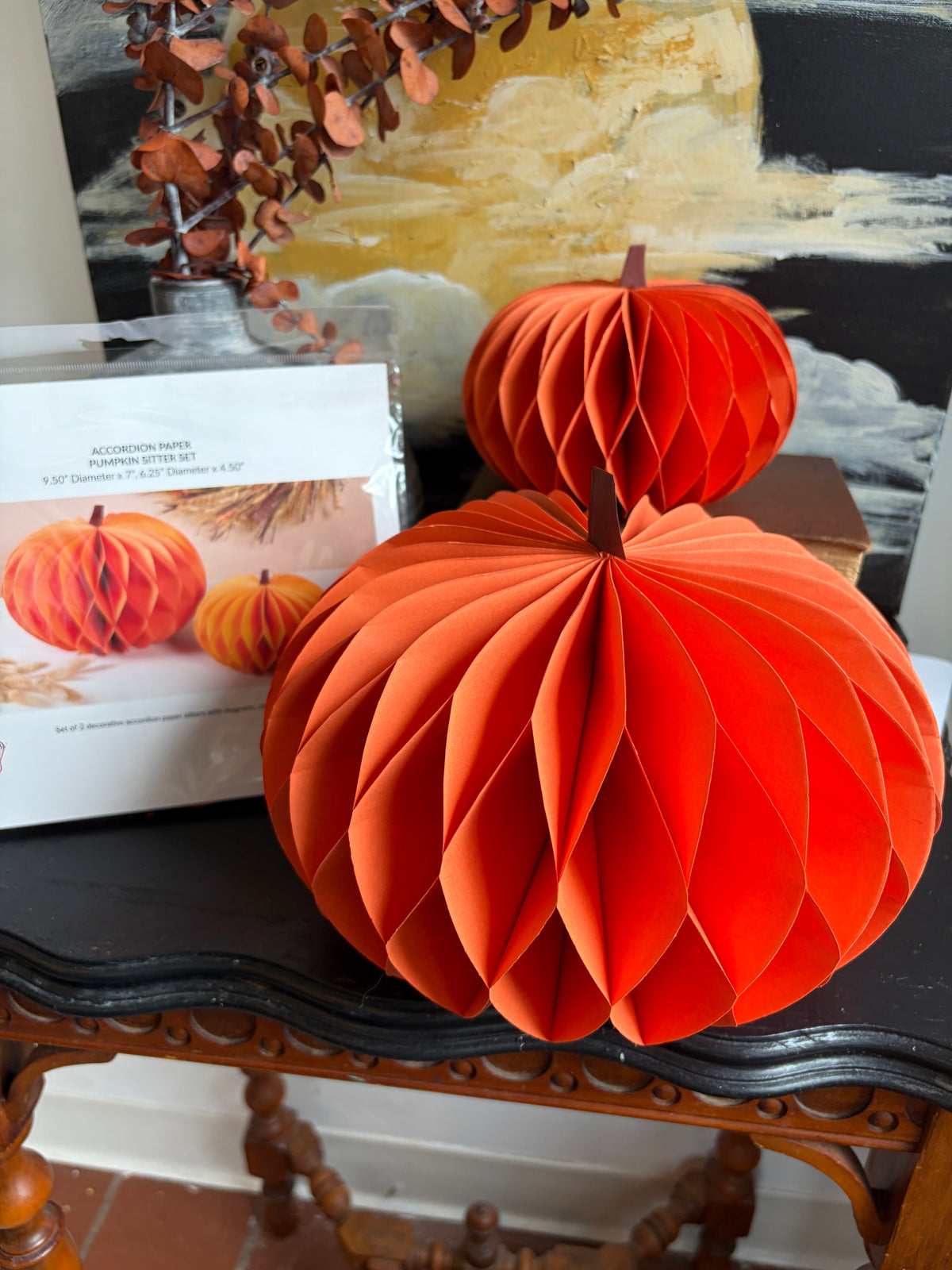 Two orange paper honeycomb pumpkin decorations displayed on a dark wooden table, with the product box visible in the background.