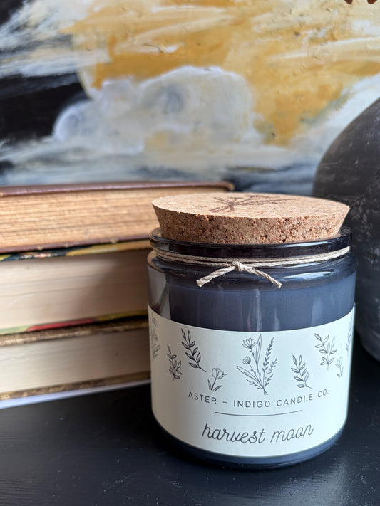 A dark gray glass jar candle with a cork lid and twine bow, featuring a white label with botanical illustrations and the text 'ASTER + INDIGO CANDLE CO.' and 'harvest moon'.