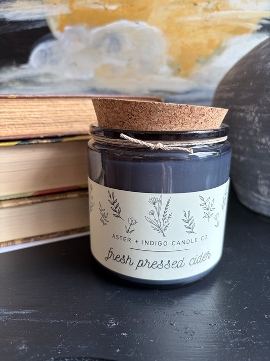 A glass jar candle with a cork lid and twine bow sits on a dark tabletop next to stacked books. The candle label reads 'ASTER + INDIGO CANDLE CO.