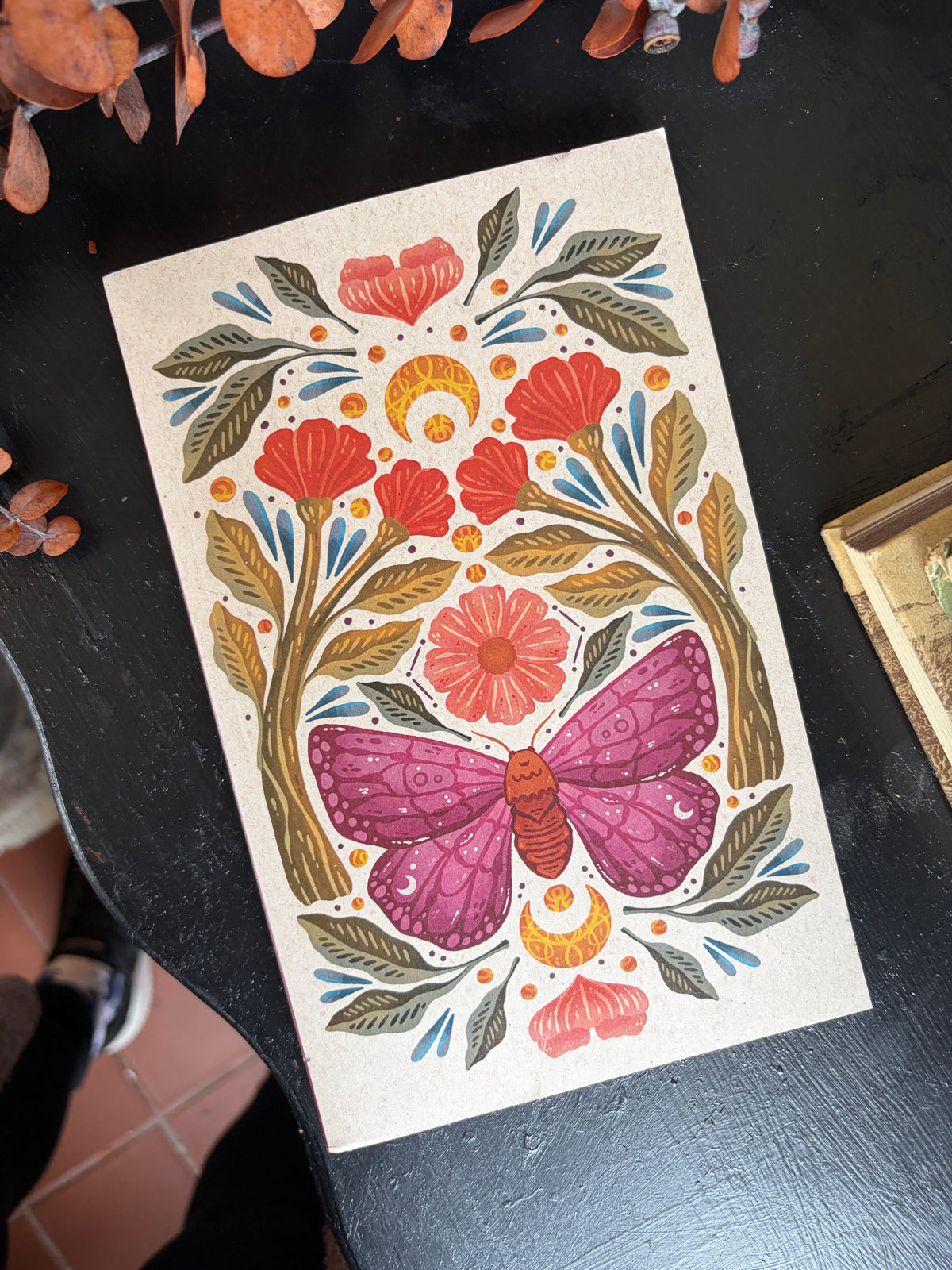 A rectangular art print or card featuring a folk-art style illustration of a pink butterfly surrounded by red flowers, green leaves, and gold crescent moons on an off-white background, photographed on a dark tabletop.