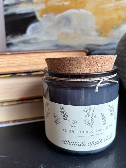A glass jar candle with a dark charcoal wax body, a cork lid, and a cream label reading 'ASTER + INDIGO CANDLE CO' and 'caramel apple chai'. It sits on a dark tabletop with stacked books and a blurred painting in the background.