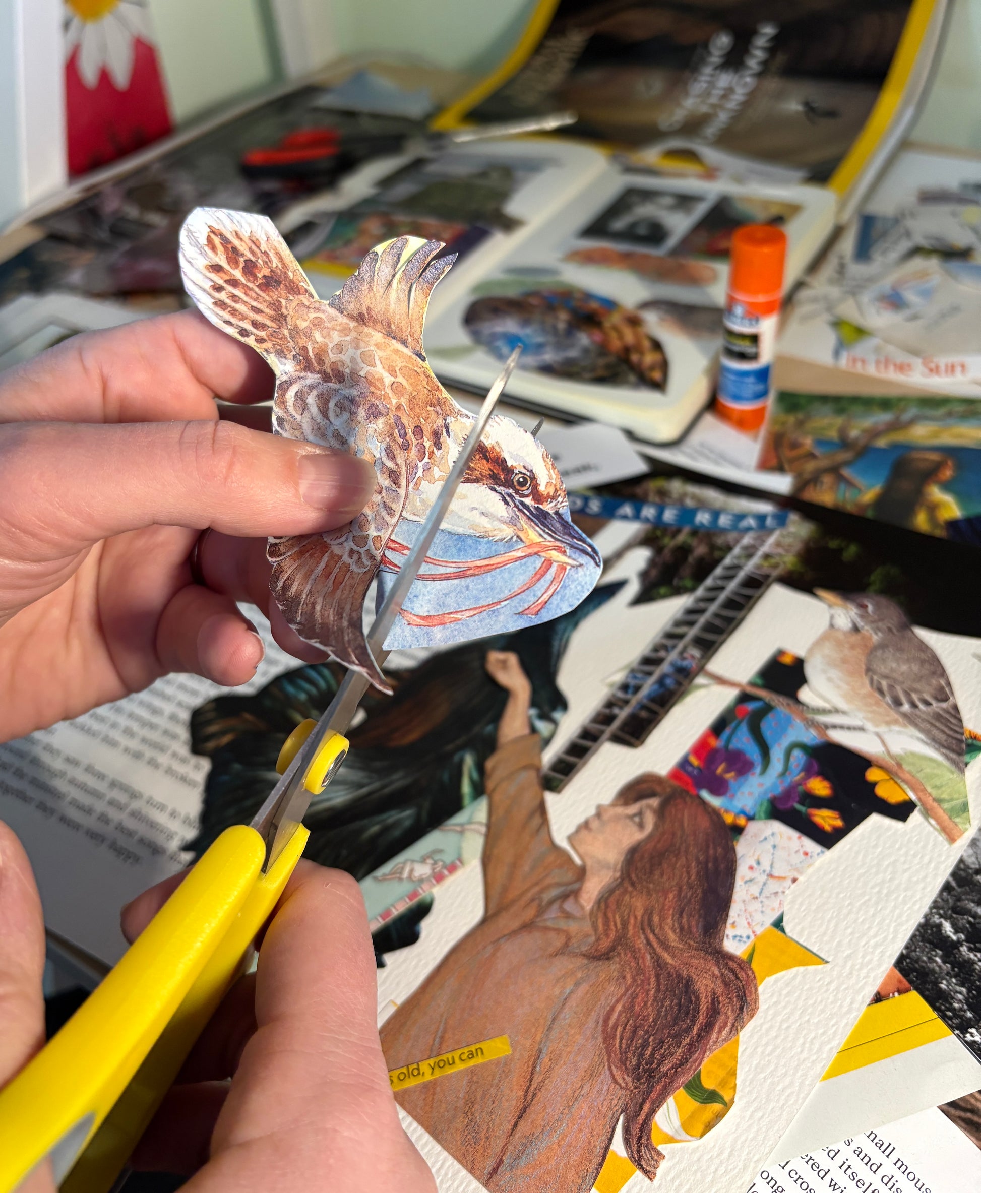Person cutting out a paper cutout of a bird from a magazine collage with collage elements  on the table underneath like birds and people.