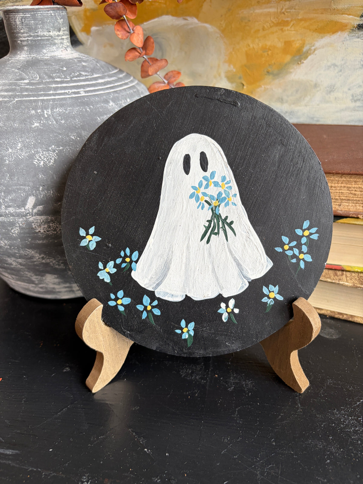 A round black slate plaque on a small wooden stand, painted with a white ghost holding blue and yellow flowers, surrounded by small blue and white flowers. The plaque is displayed on a dark surface with a rustic gray ceramic vase and books in the background.