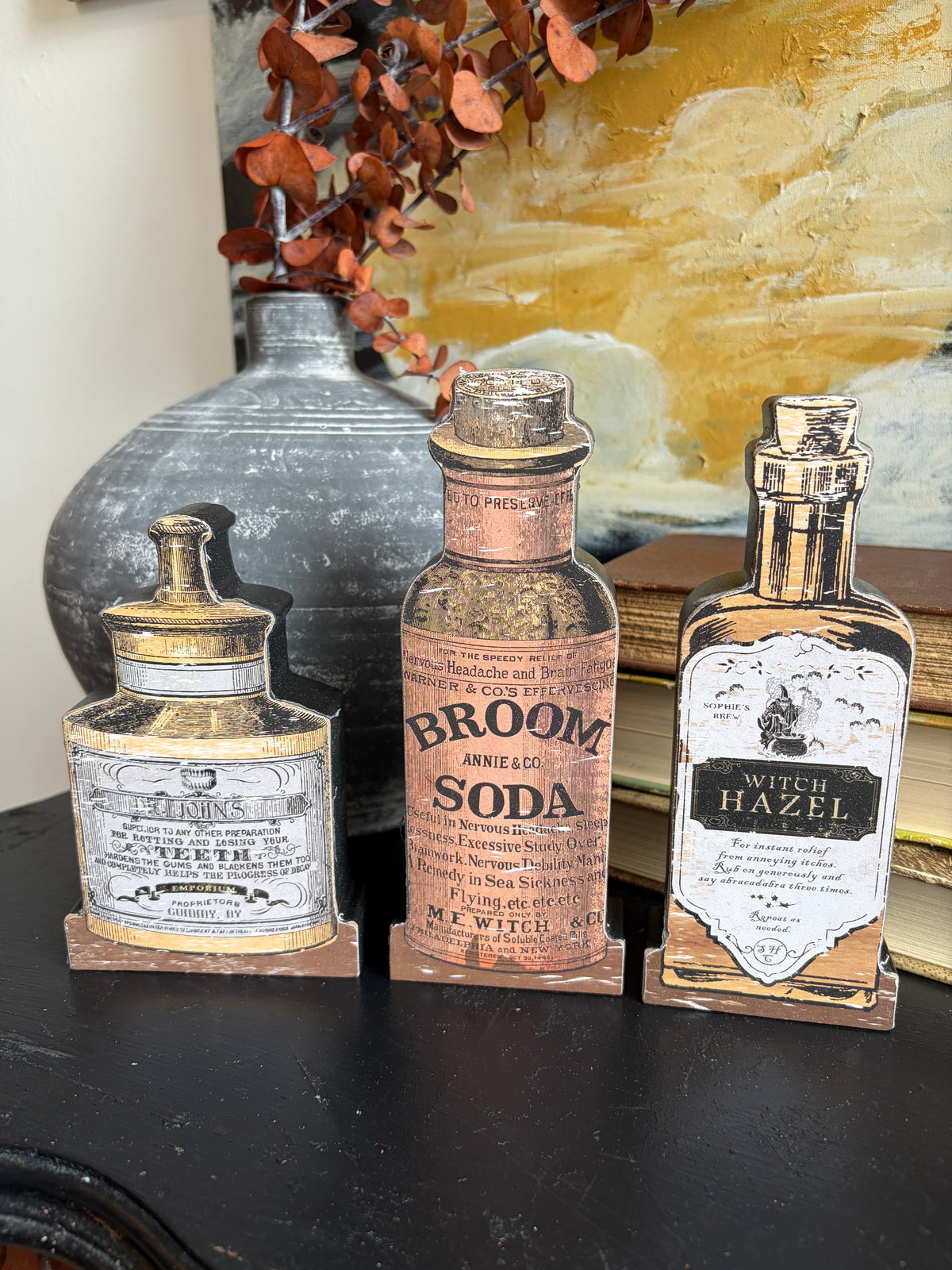 Three vintage-style decorative bottle cutouts displayed on a dark tabletop, featuring labels such as 'Broom Angel' and 'Witch Hazel,' with a gray vase and warm-toned artwork in the background.