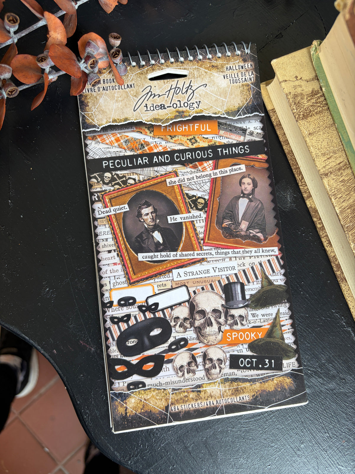 A spiral-bound Tim Holtz Layering Journal page featuring a spooky collage design with portraits, masks, skulls, and text like 'Peculiar and Curious Things' and 'Nighty.' It is photographed on a dark tabletop next to books and decorative branches.