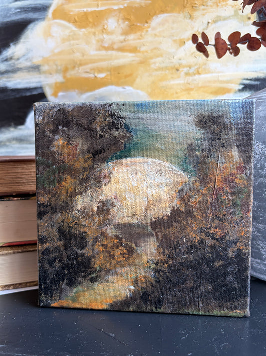 A small square canvas painting depicting a moonlit forest scene with dark trees and a glowing pale moon, displayed on a dark surface with books and another artwork in the background.