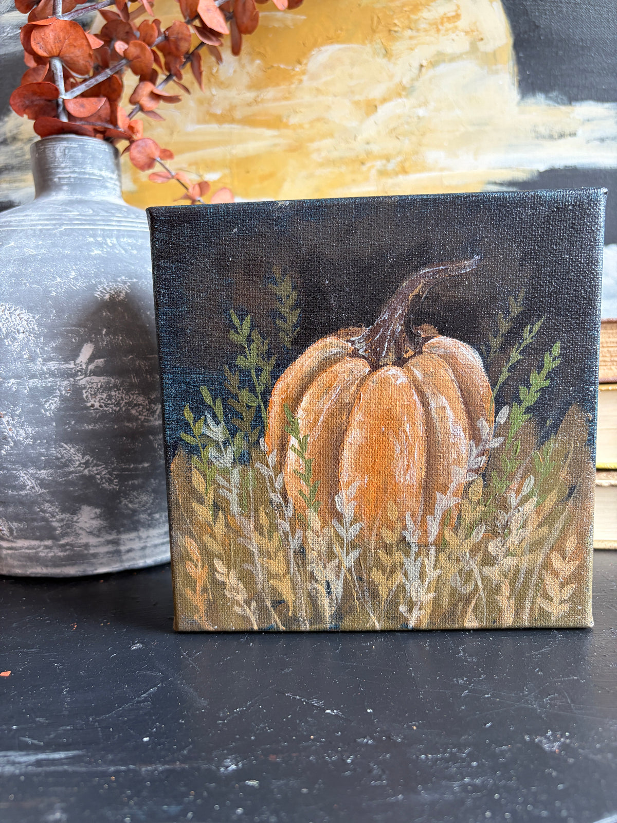 A small square canvas painting of an orange pumpkin in a field of tall grass, displayed on a dark tabletop next to a gray ceramic vase with dried orange leaves.