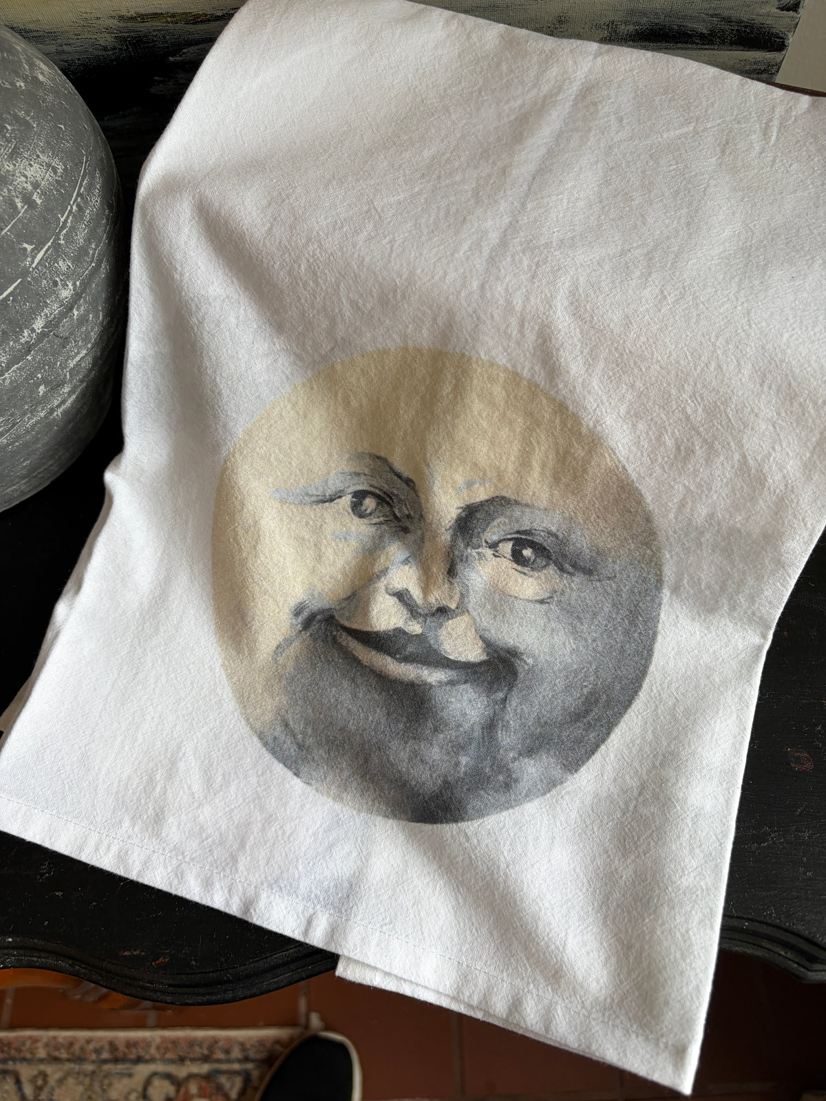 A white fabric tote bag or cloth bag laid on a dark table, featuring a circular printed illustration of a smiling moon face in beige, gray, and black tones.