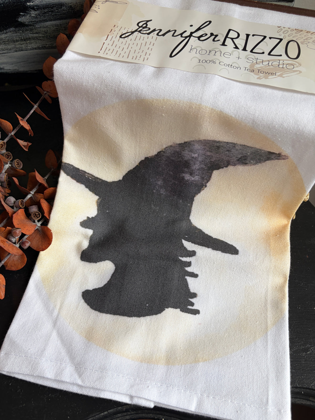 A white cotton tea towel with a black silhouette of a witch flying on a broomstick over a large beige circle, featuring a paper header that reads 'Jennifer RIZZO home + studio'.
