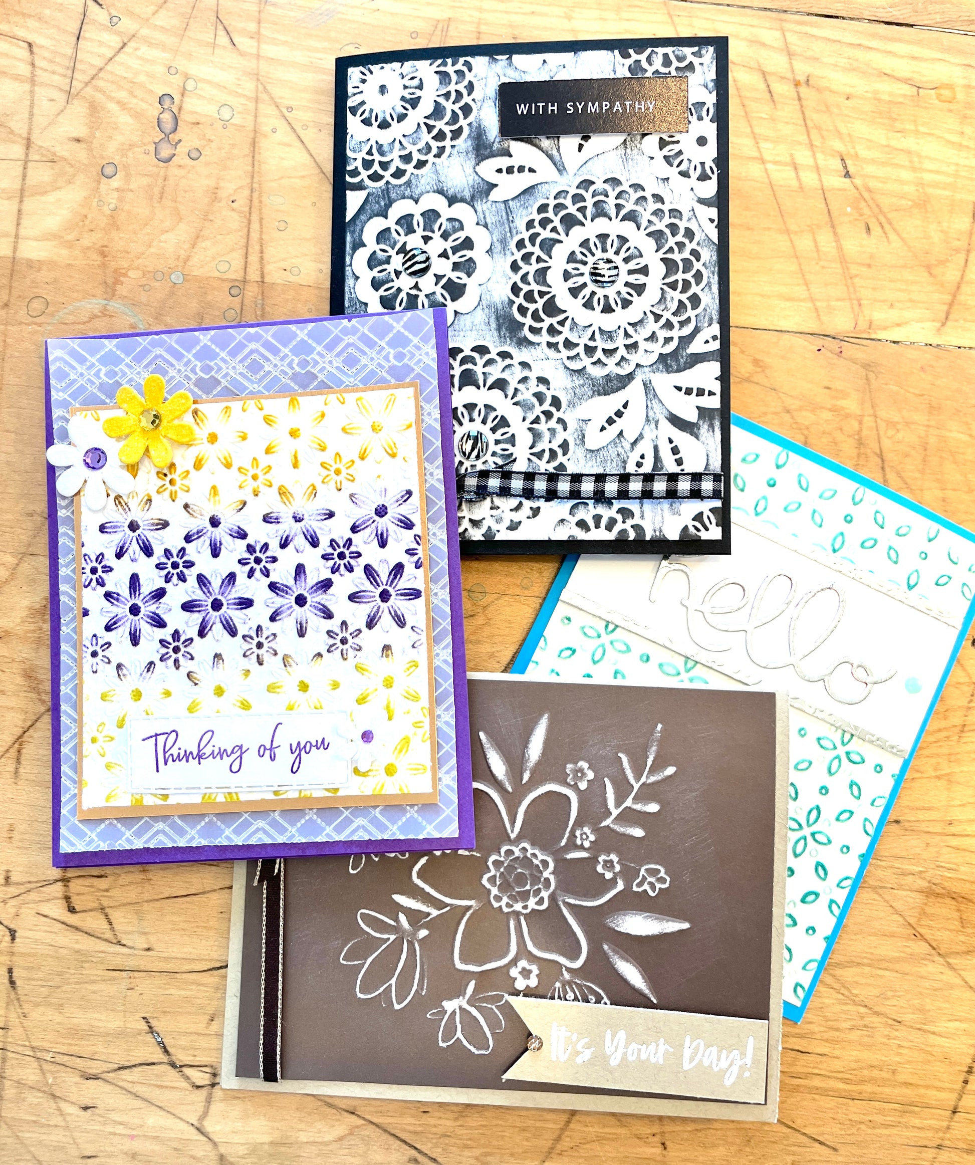Four greeting cards with floral designs on a wooden surface. It shows a set of embossed greeting cards. One says hello, others say with sympathy, it's your day and thinking of you with a yellow flower.