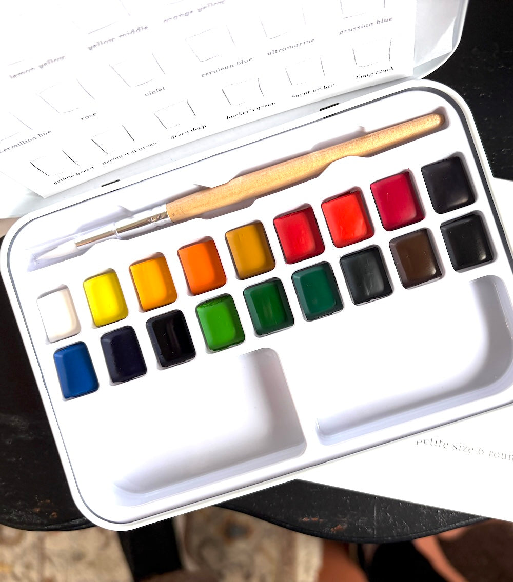 A 16-color watercolor paint set in a white metal tin, shown open with a wooden-handled paintbrush resting inside. The paint pans are arranged in two rows, displaying a full spectrum of colors including yellow, orange, green, blue, red, black, and white.