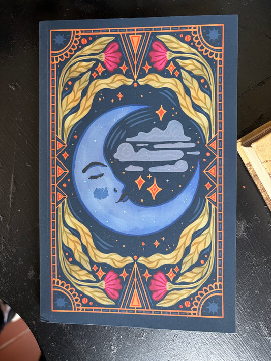 A square art print or card featuring a stylized blue crescent moon face with clouds and stars, surrounded by ornate gold and pink decorative borders on a dark navy background, photographed on a dark tabletop.
