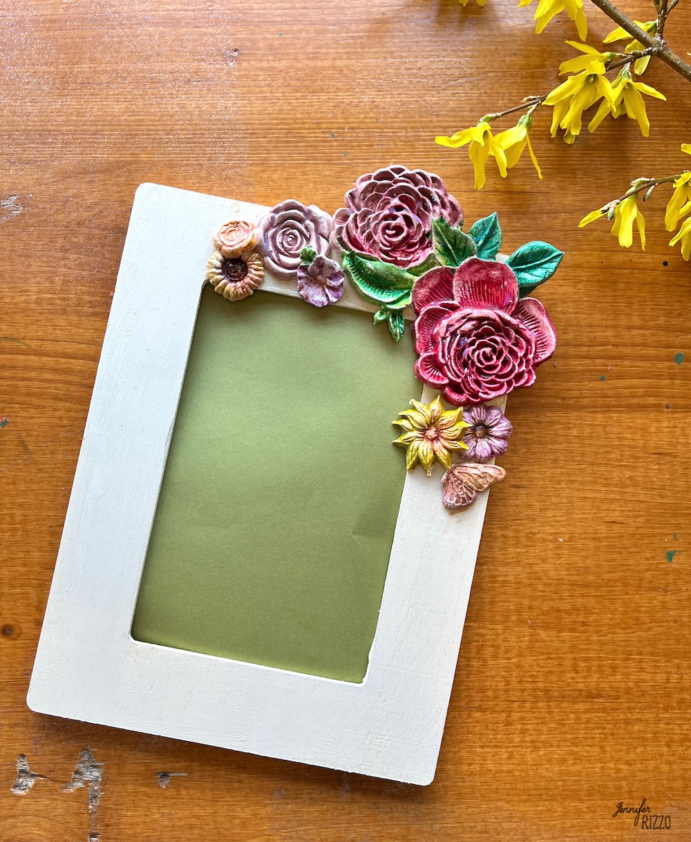 Image of a picture frame on a wood surface with colorful flowers glued on it with real yellow flowers next to it.
