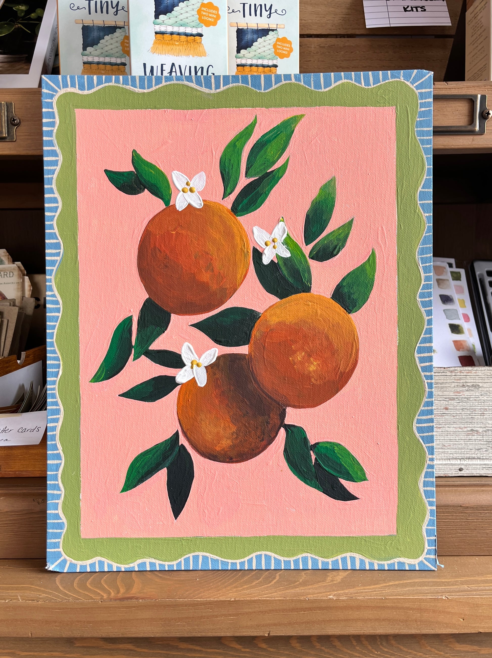 Painting of oranges and flowers in acrylic paint with green leaves on a pink background. The edges are painted in a wavy green and blue pattern that looks like a frame. 