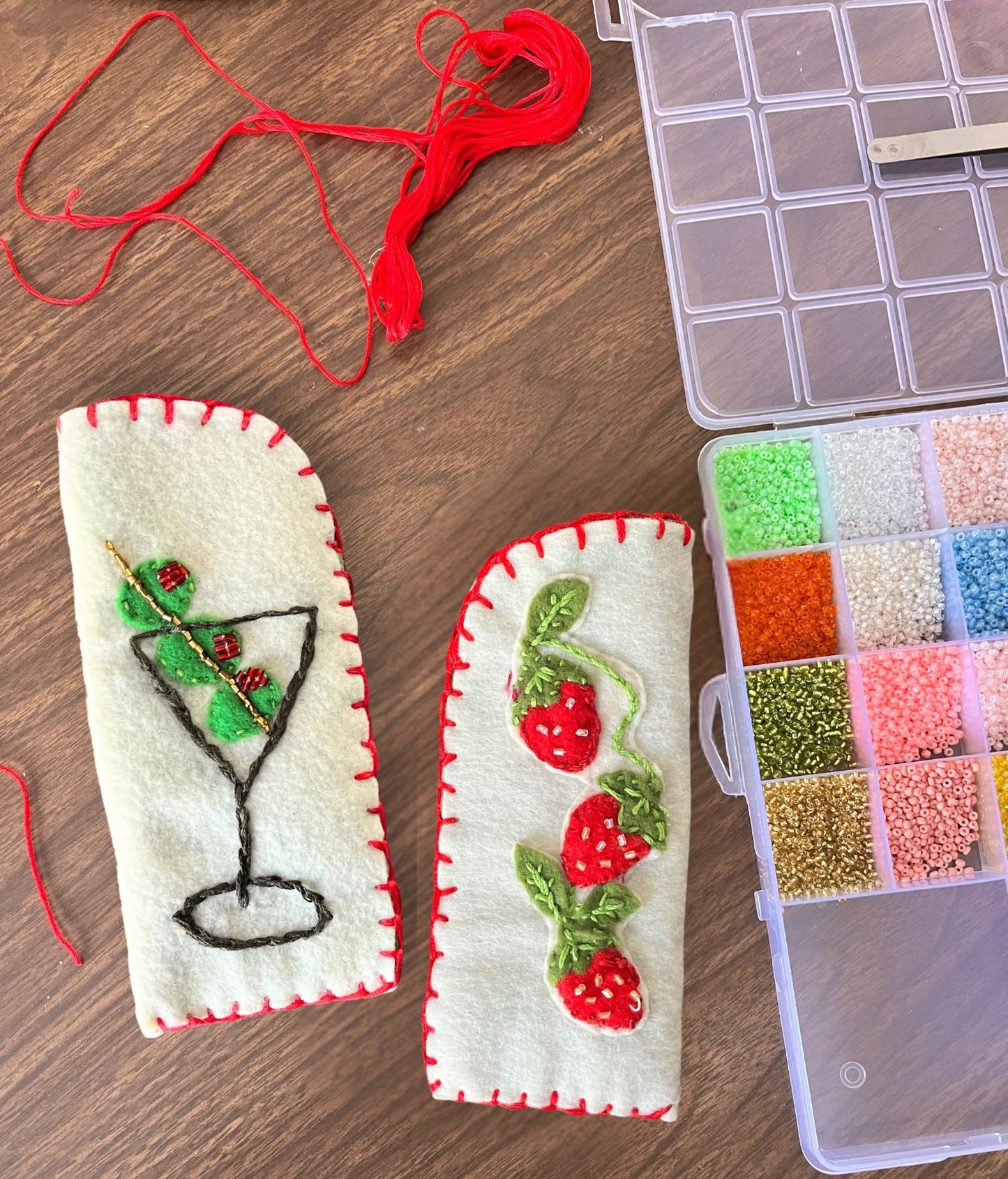 Two embroidered eye glass cases with a martini glass and strawberry design on a wooden surface with embroidery supplies.