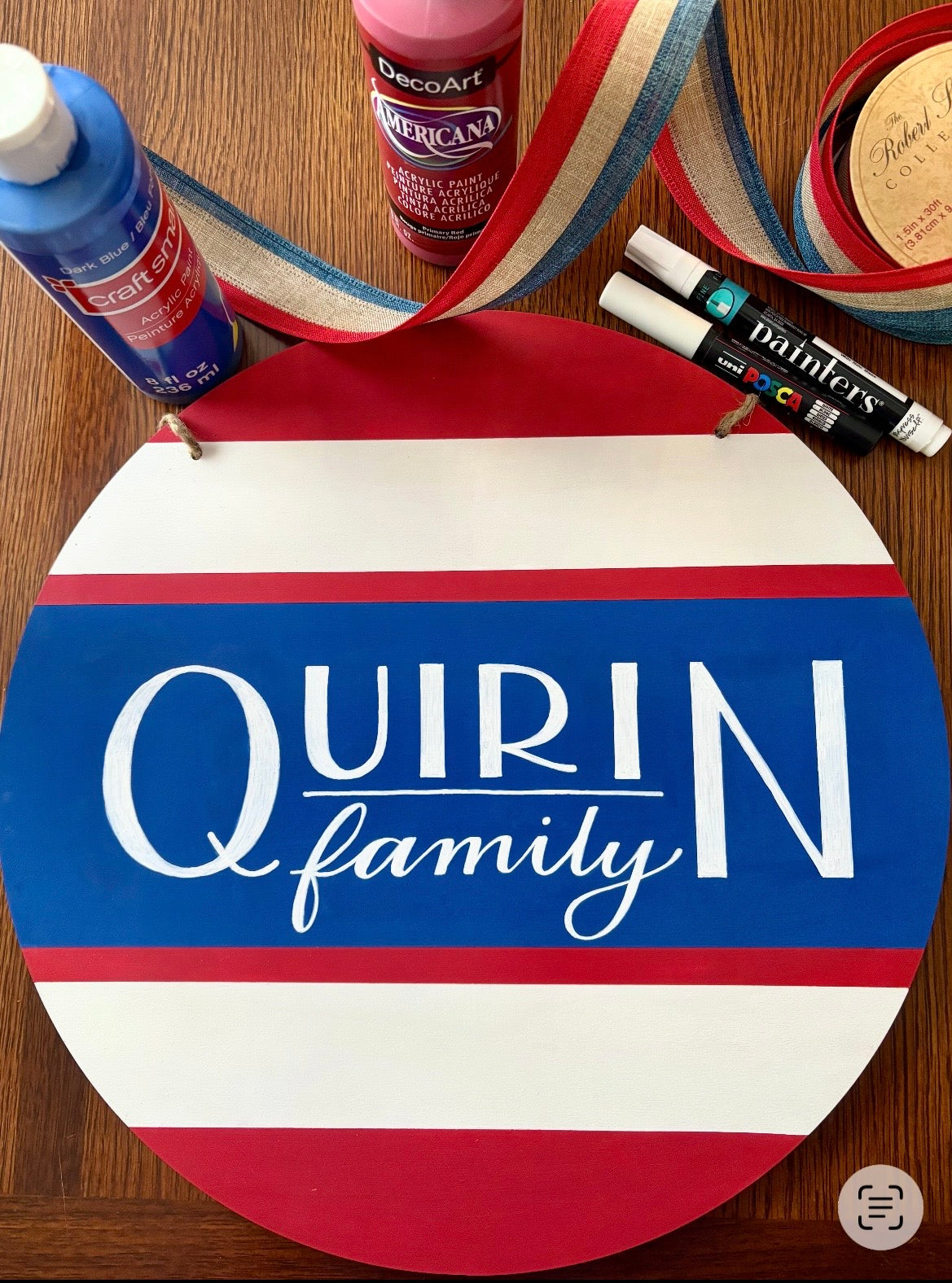 Round wooden sign with 'Quirin Family' text, surrounded by craft supplies on a wooden surface.