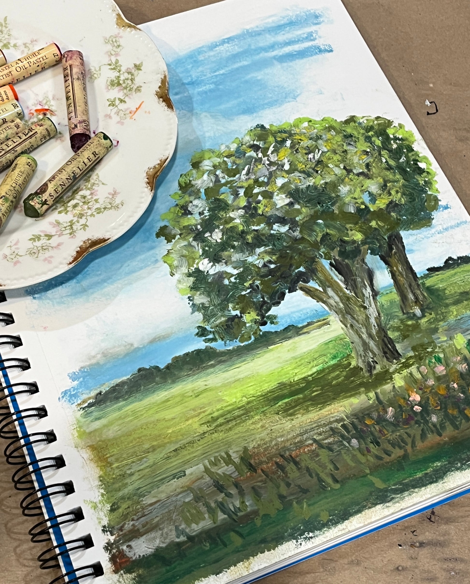 Painting of a landscape with trees and grass on a sketchbook page with a vintage dish of oil pastels to the right