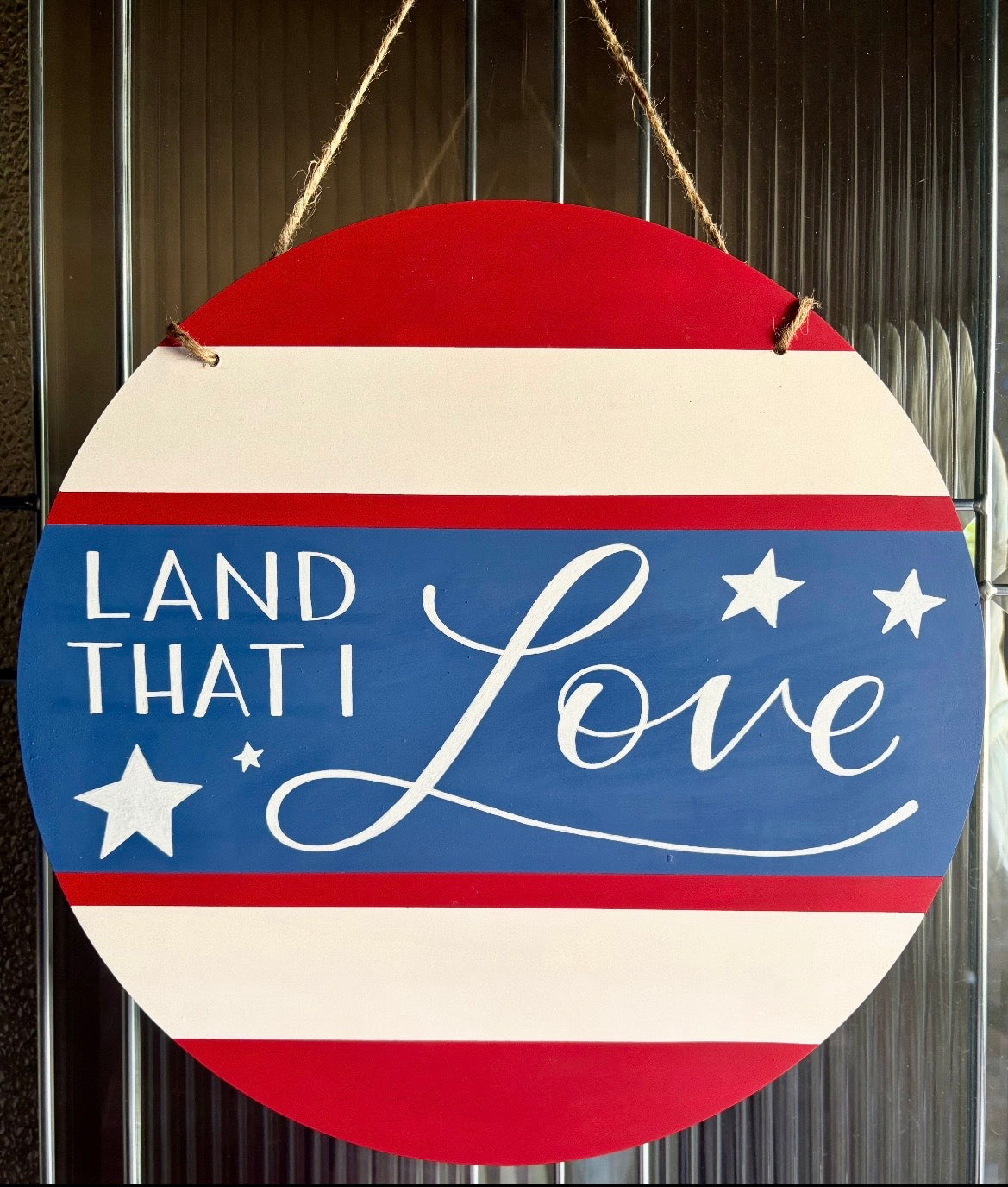 Round decorative sign with red, white, and blue stripes and 'Land That I Love' text.