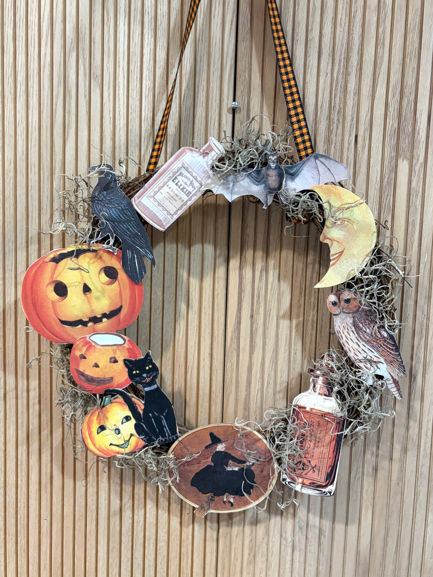 A Halloween-themed decorative wreath hanging on a light wood paneled wall. The wreath is made of natural jute and adorned with printed autumn motifs including jack-o'-lanterns, a black cat, a witch silhouette, a bat, a owl, a crescent moon, a crow, and vintage-style bottles, with a black-and-orange plaid hanging ribbon at the top.